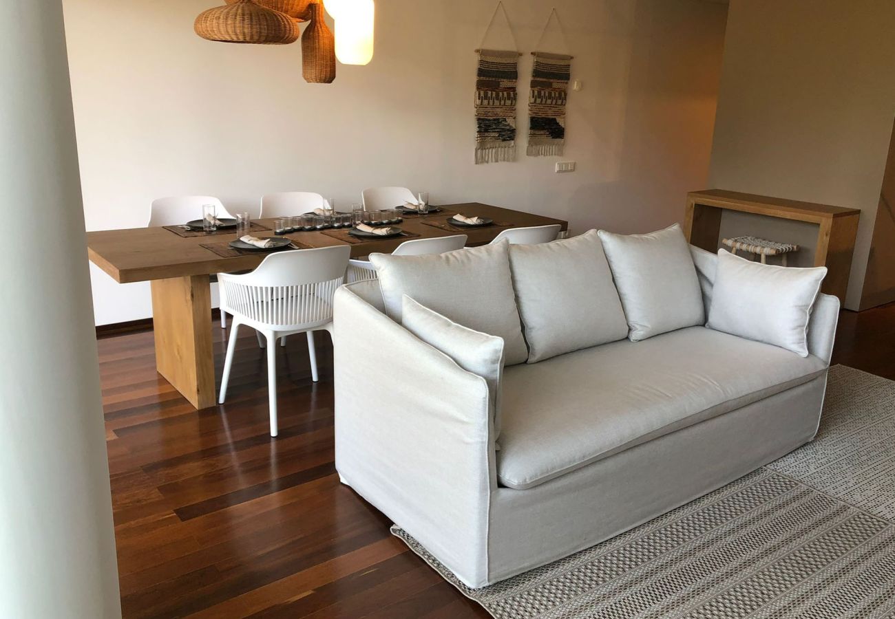 Wooden dining table with white chairs and pendant lighting, next to an elegant sofa in a cozy and modern space.