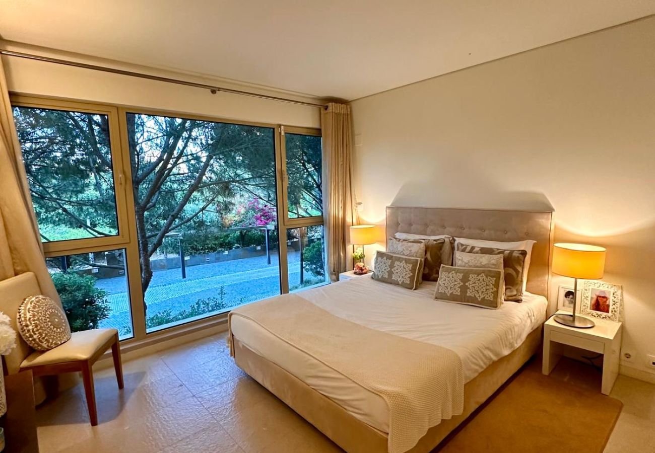Spacious bedroom featuring a double bed, upholstered headboard, and soft lighting. Large glass windows provide a view of the garden and allow natural 