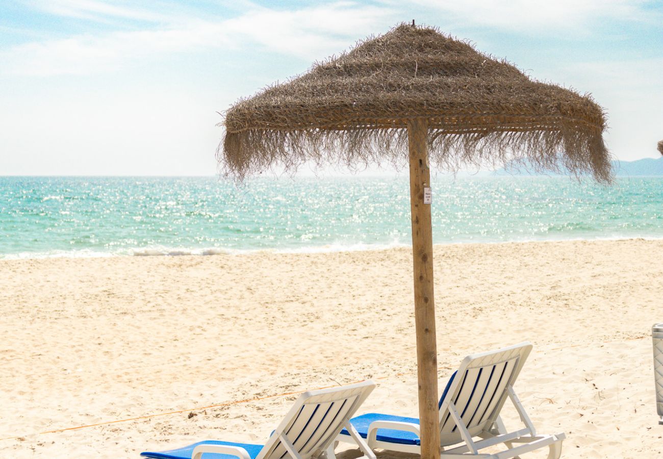 View of a white sand beach with a straw umbrella in the center and two blue lounge chairs facing the crystal-clear sea under a blue sky with some clou