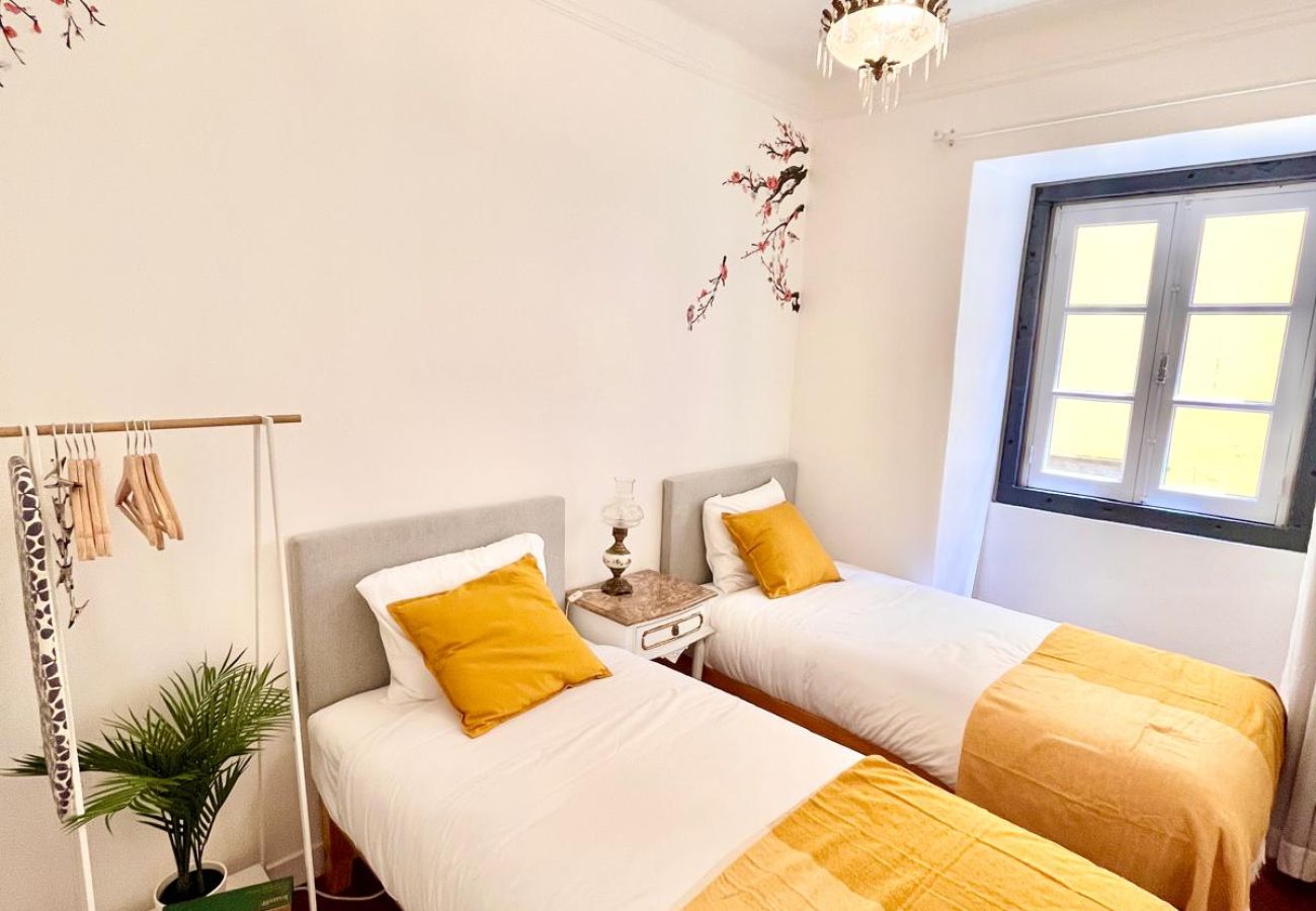 Bright room with two beds, yellow cushions, large window, and a decorative tree detail on the wall.