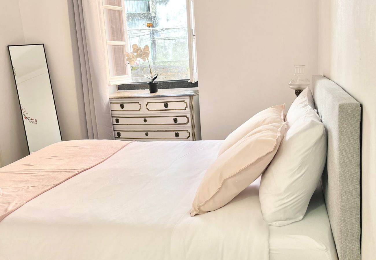Bedroom with double bed, upholstered headboard, mirrored nightstand, and open window with light curtains.