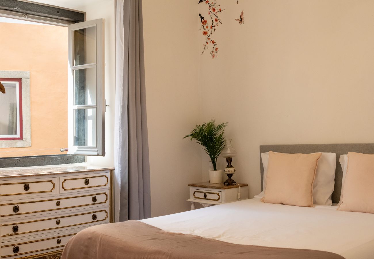 Bedroom with double bed, upholstered headboard, mirrored nightstand, and open window with light curtains.