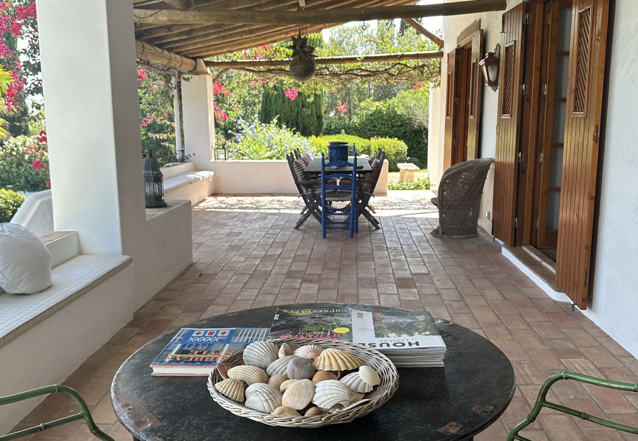 Cozy terrace with a wooden roof, round table decorated with seashells, cushioned chairs, and a view of the blooming garden.