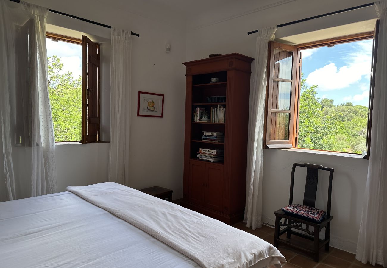 Cozy bedroom with a double bed, open wooden windows with flowing white curtains, a nature view, a classic wooden wardrobe, and a decorative chair.