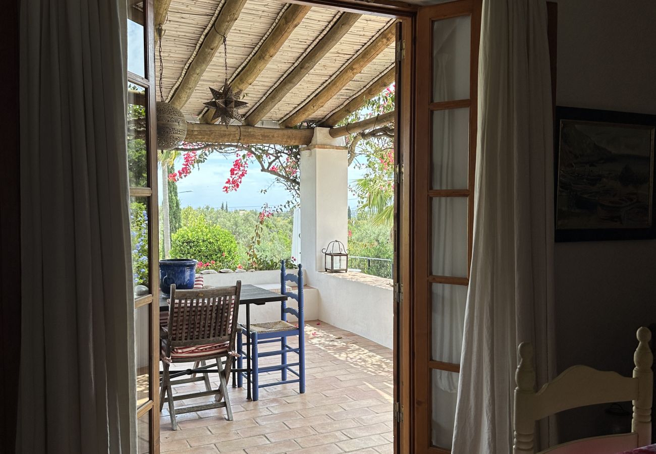 Wooden door with open curtains leading to a covered terrace with a table and chairs, overlooking a flowered garden and landscape.