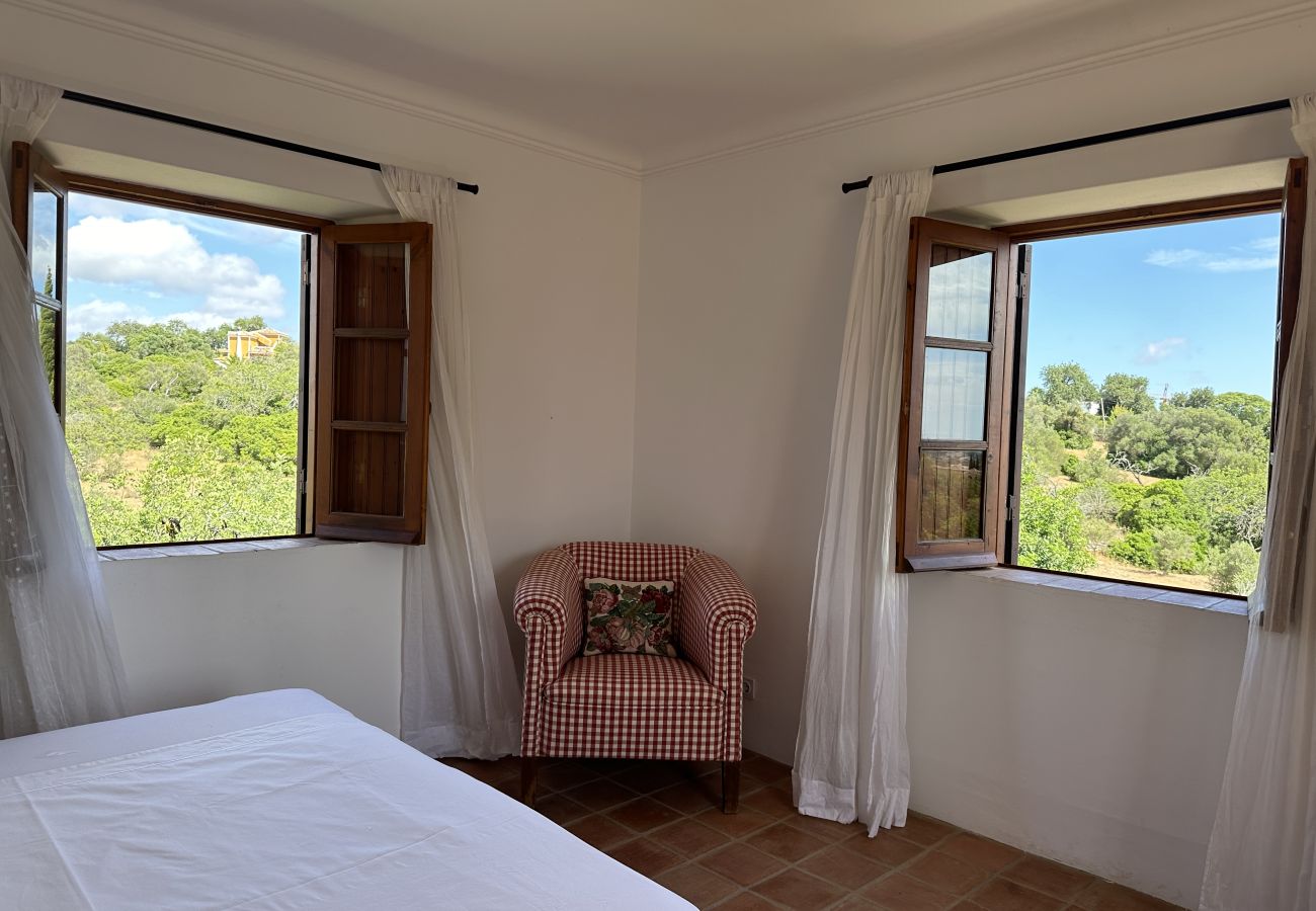 Cozy bedroom with a double bed, a comfortable armchair, and open windows revealing a stunning green landscape.