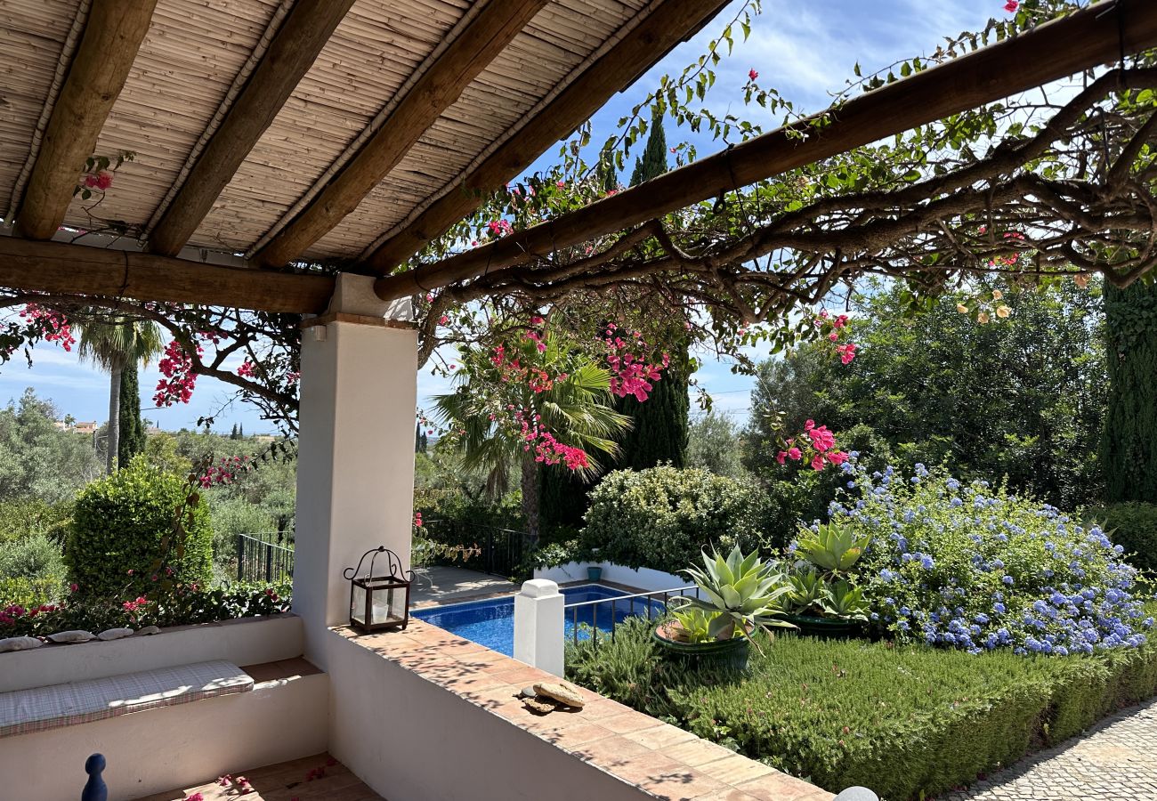 Covered terrace with blooming vines, a decorative lantern, and a view of a lush garden with a pool in the background.