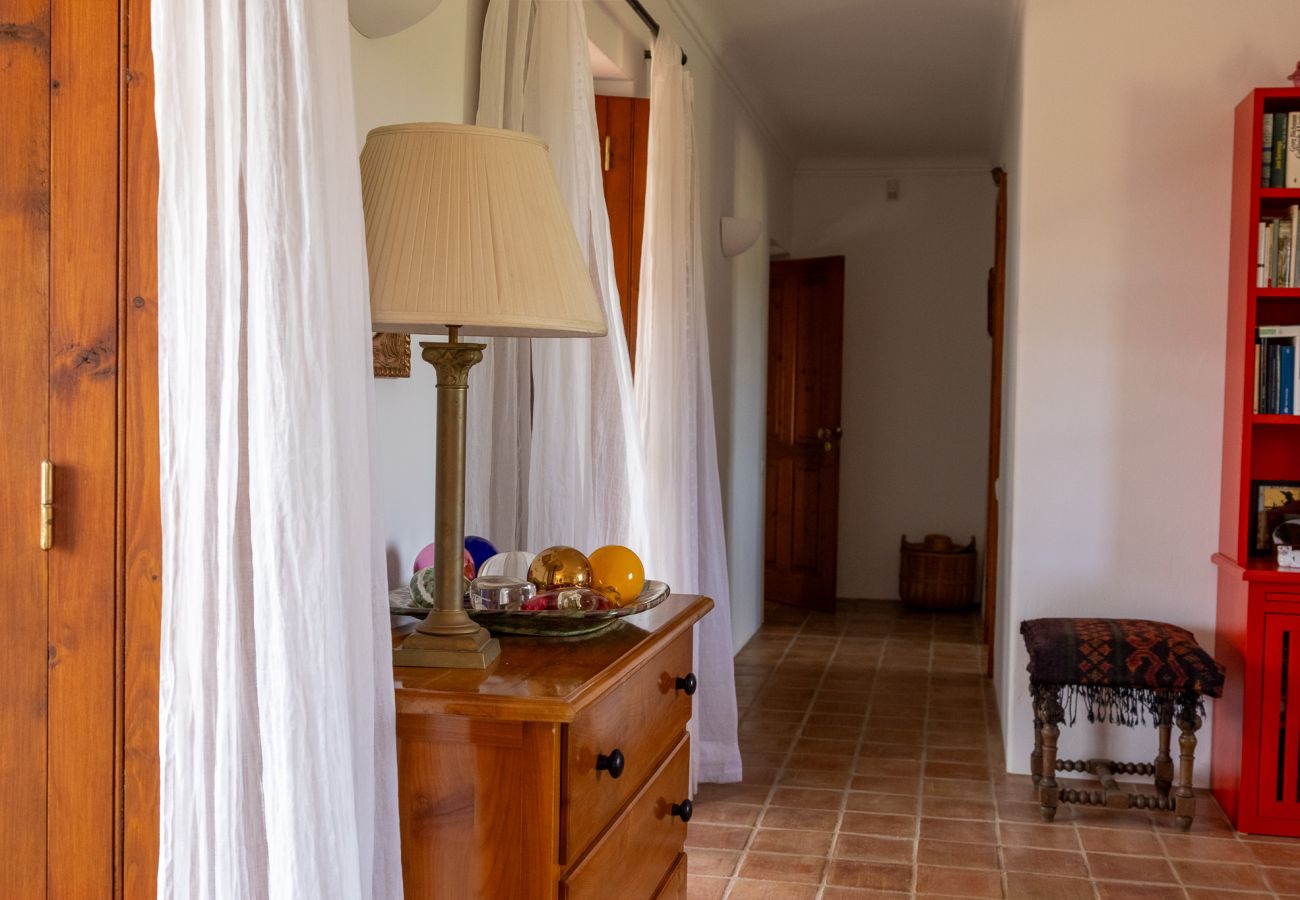 Bright hallway featuring a wooden dresser, classic lamp, white curtains, and a red bookshelf filled with books. In the background, an open door and a 