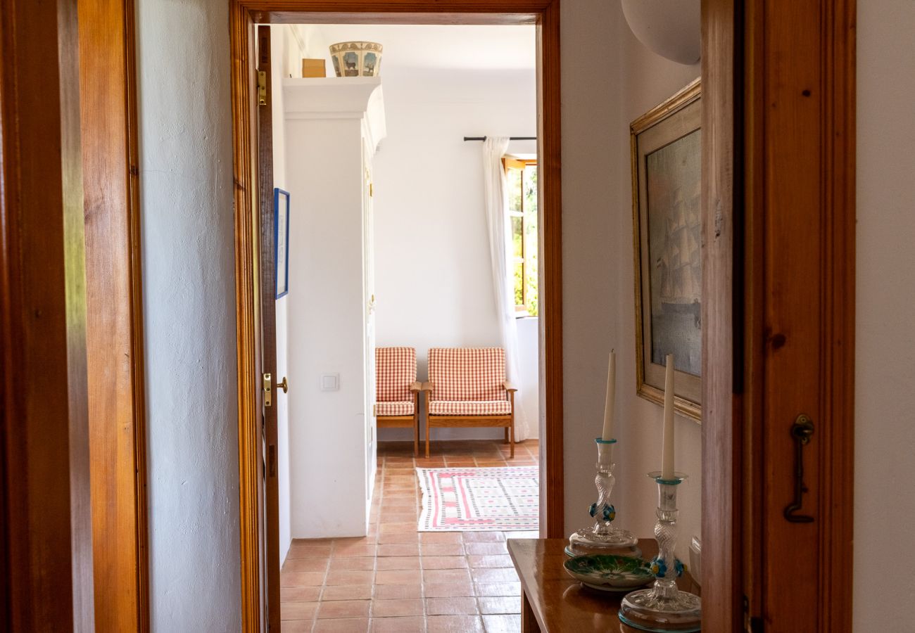 Hallway with wooden furniture, decorative candles, and a painting on the wall, leading to a bright bedroom with armchairs and a colorful rug.