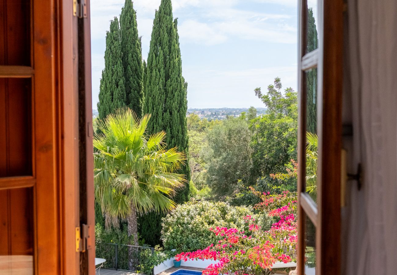 Open wooden window revealing a lush garden with a blue pool and vibrant greenery in the background.