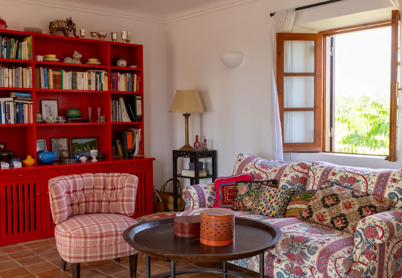 Cozy living room featuring a red bookshelf filled with books and decorative items, a patterned sofa with colorful cushions, and open wooden windows ov
