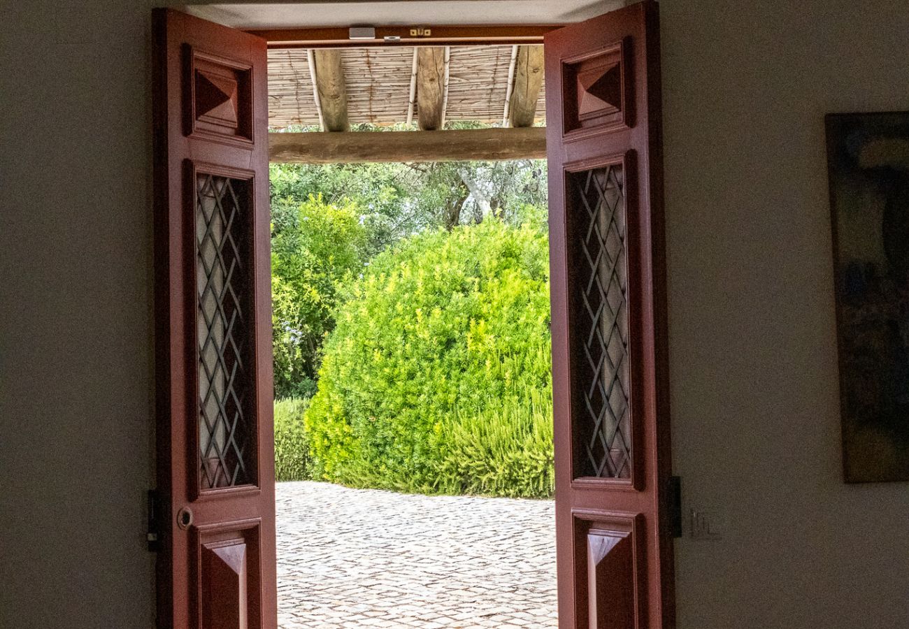  Open double wooden door revealing an outdoor courtyard with a stone floor, lush greenery, and rustic architectural details.