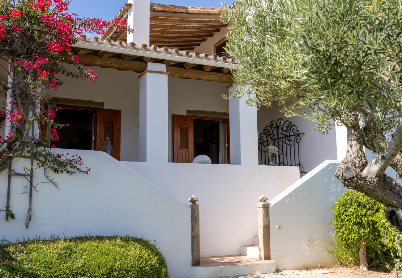 White façade of the villa in Almancil, featuring a wooden-covered porch, blooming bougainvillea, and a lush green garden.