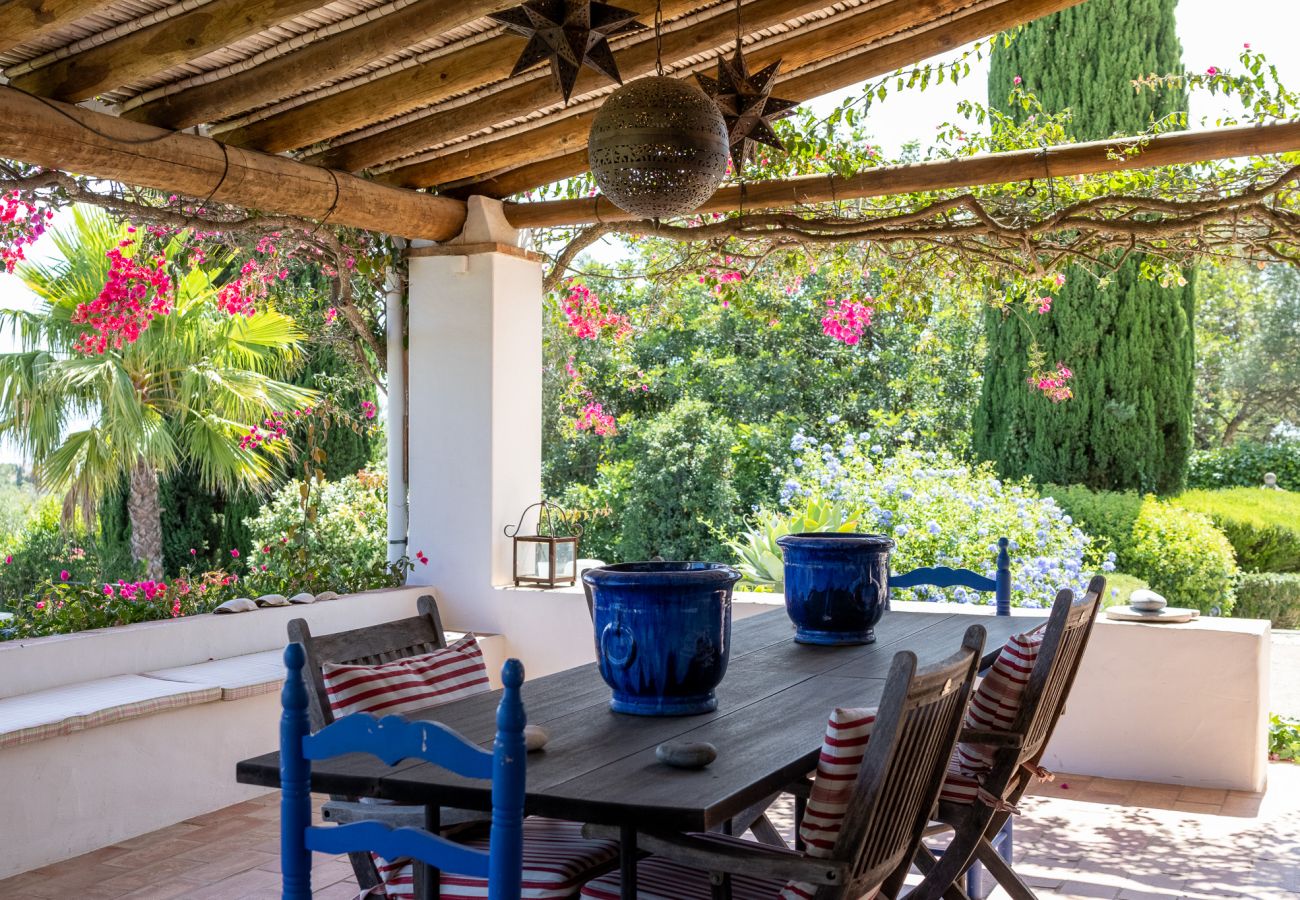 Covered terrace with a dining table, rustic decor, and a view of the lush garden in Almancil.