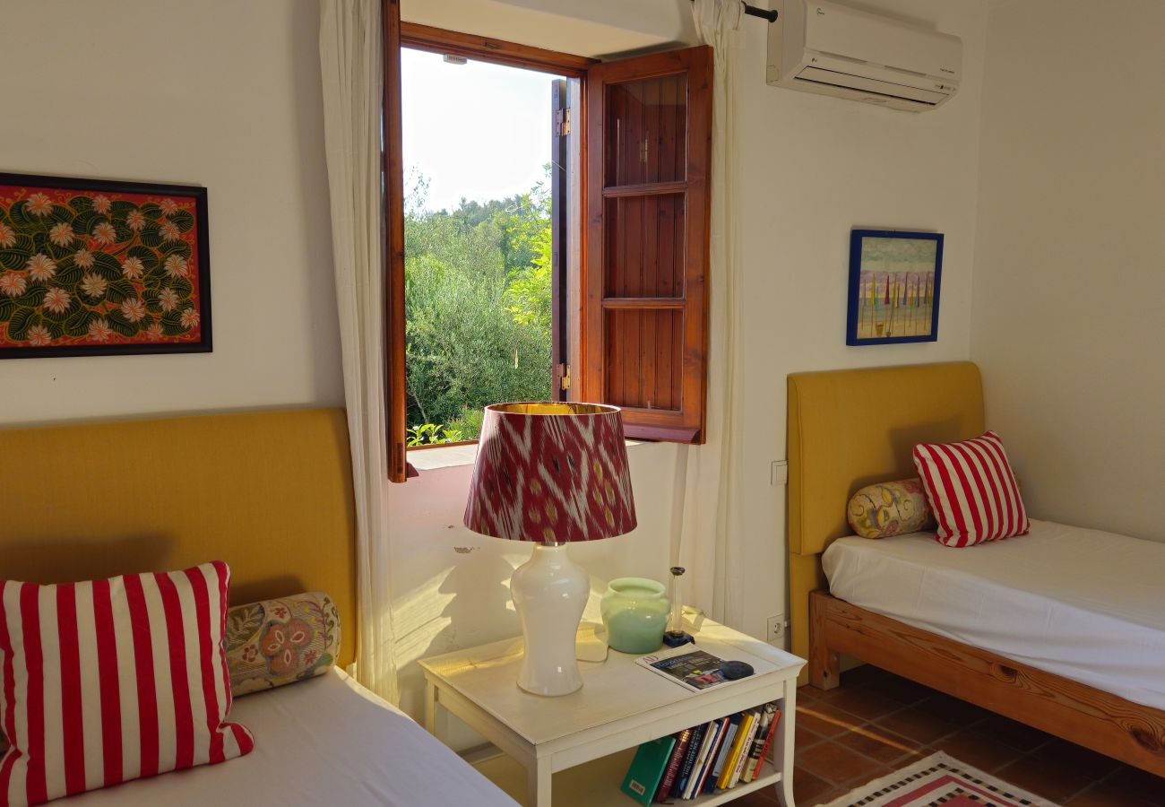 Bedroom with two single beds, yellow headboards, striped pillows, decorative lamp on a nightstand with books and objects, open windows with a view of 