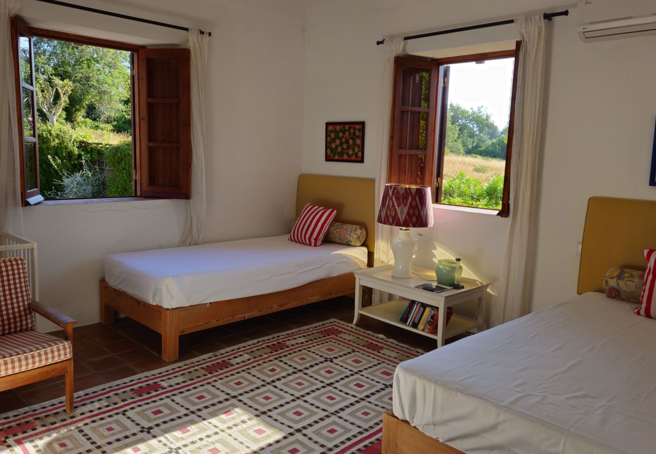 Bright bedroom featuring two single beds, rustic furniture, and open windows offering a beautiful view of the surrounding greenery. Patterned rug and 