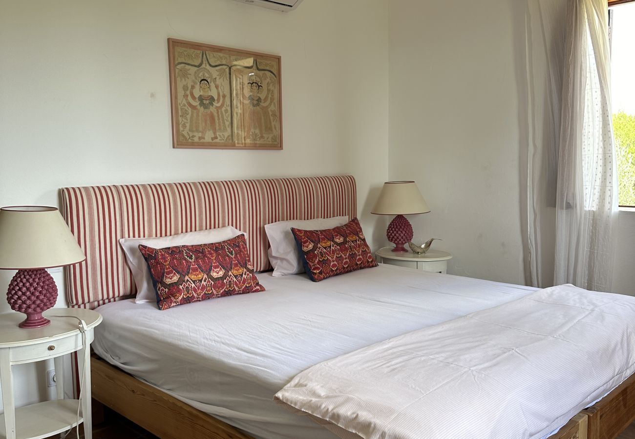 Comfortable bedroom with a double bed, striped headboard, decorative pillows, elegant bedside lamp, and an open wooden window with light white curtain