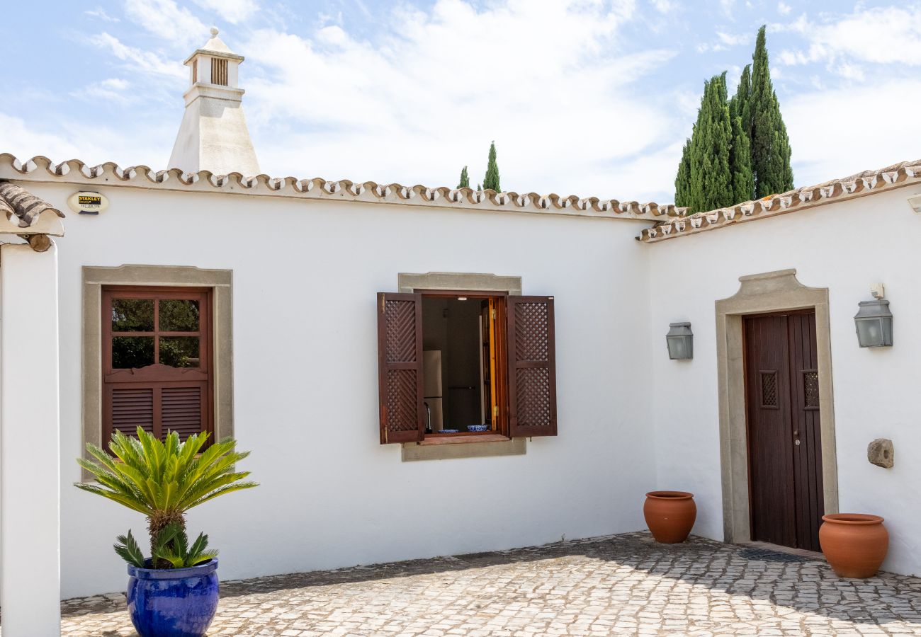 White house with traditional architecture, wooden door, shuttered windows, and a stone-paved courtyard decorated with potted plants.