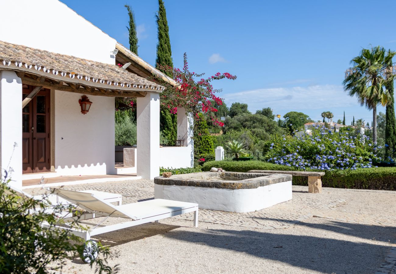 Exterior of the villa in Almancil featuring sun loungers, a rustic fountain, and a garden view.