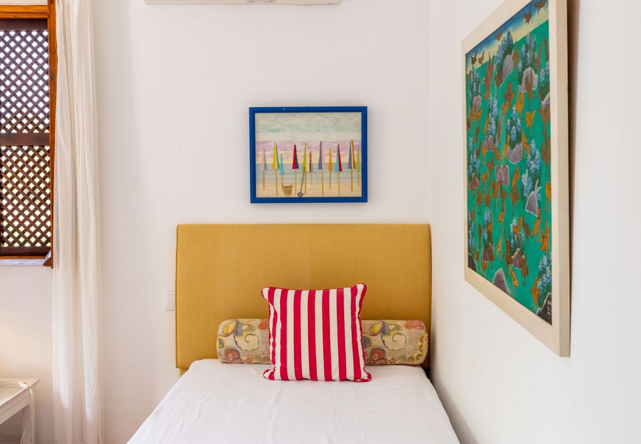 Minimalist bedroom featuring a single bed with a yellow headboard, a red and white striped pillow, colorful paintings on the wall, and a window with a