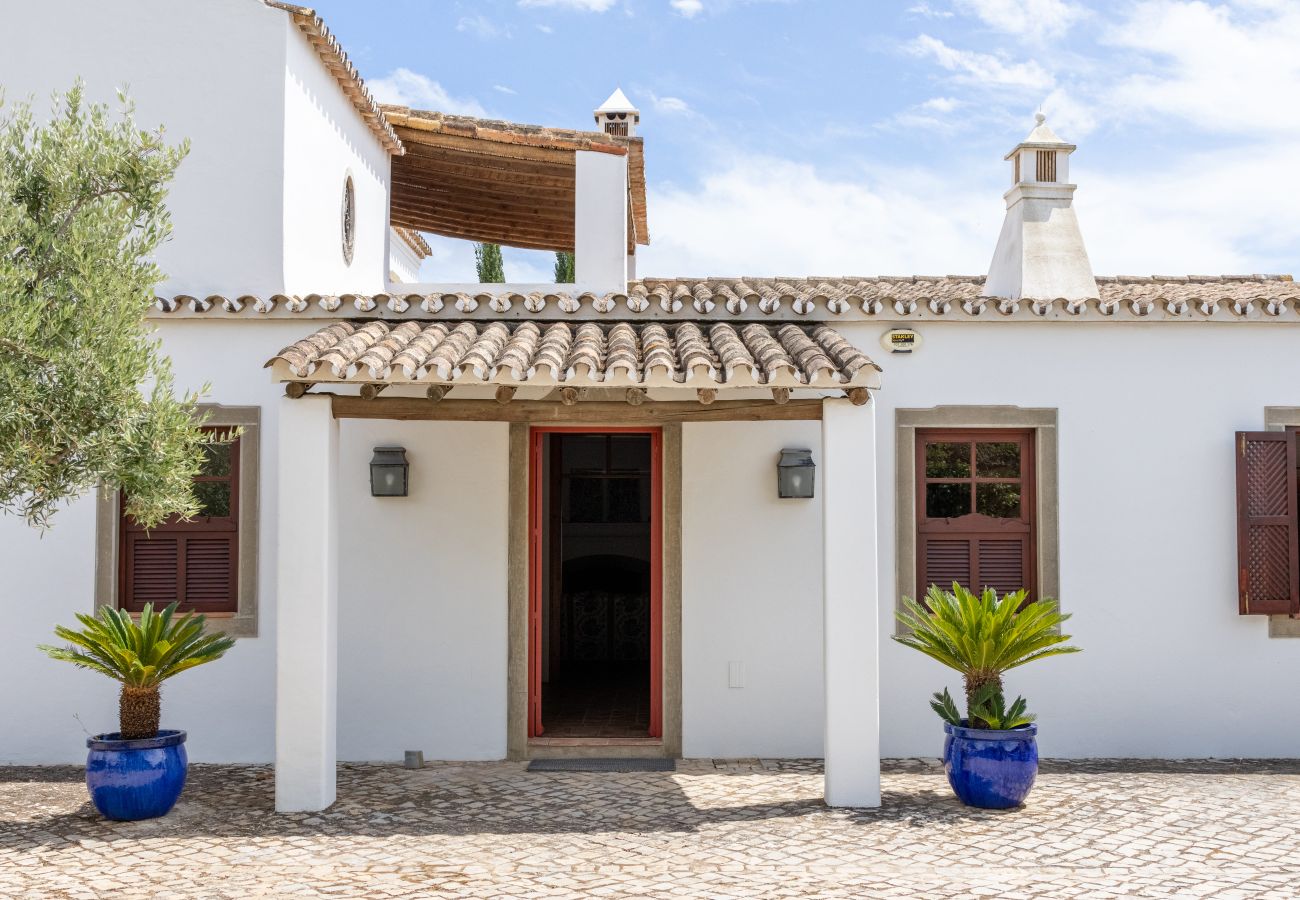 White house with traditional architecture, wooden door, shuttered windows, and a stone-paved courtyard decorated with potted plants.