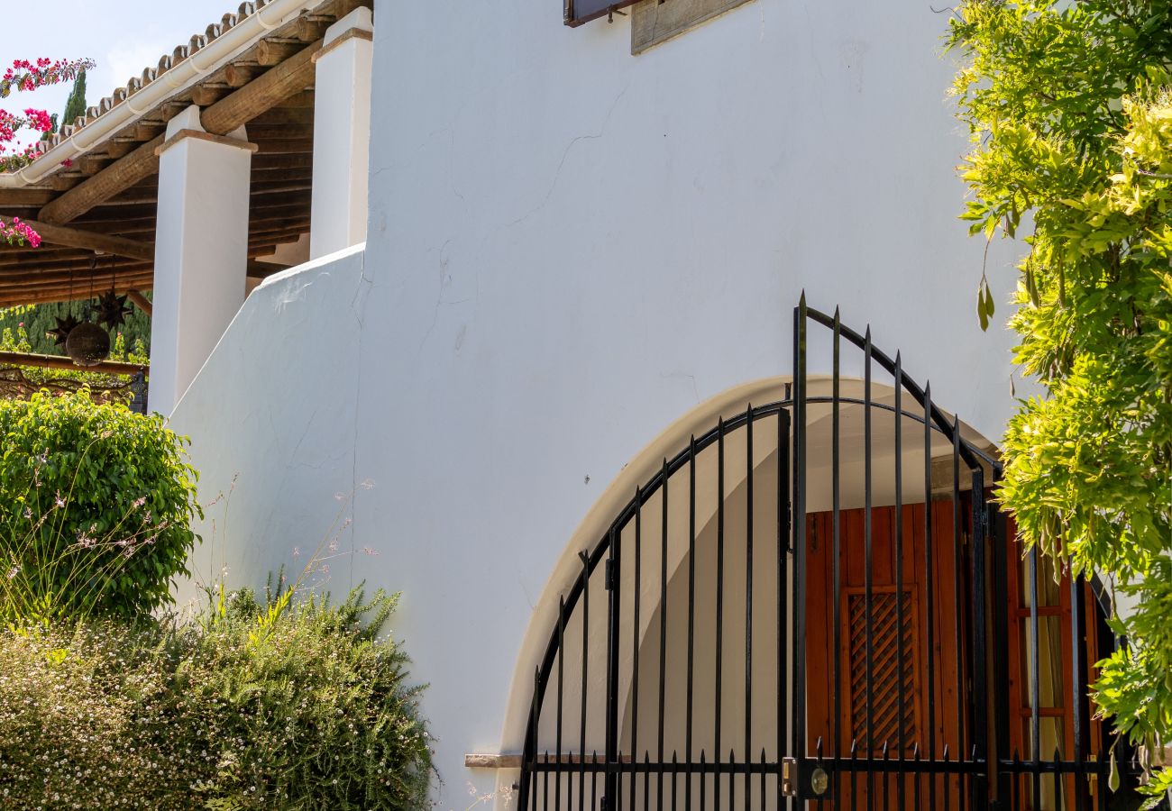 White house facade with an arched iron gate, wooden shuttered window, and lush greenery, showcasing rustic and elegant architecture.