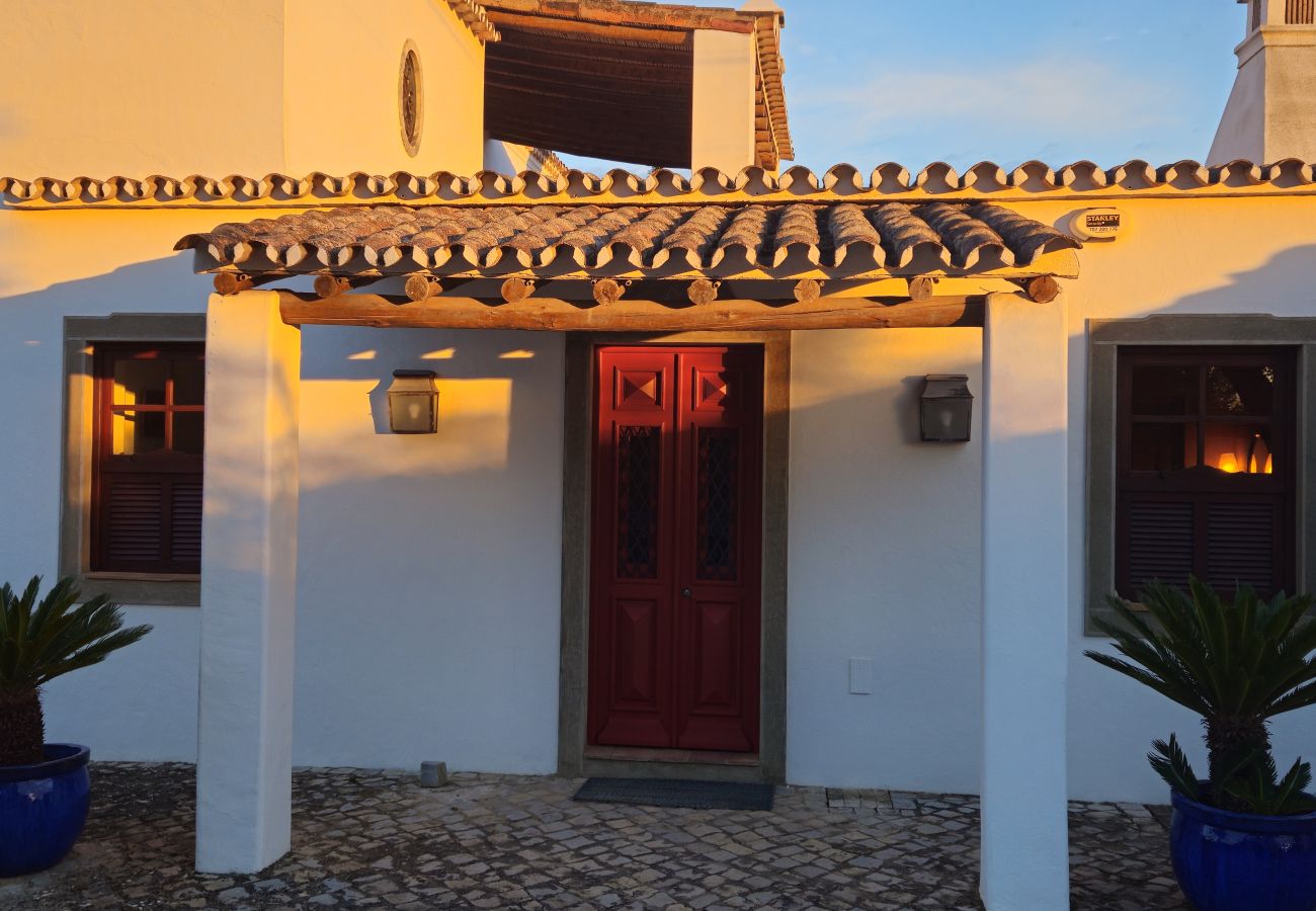 Entrance of a white house with a red door, under a tiled porch, illuminated by the golden light of the sunset.