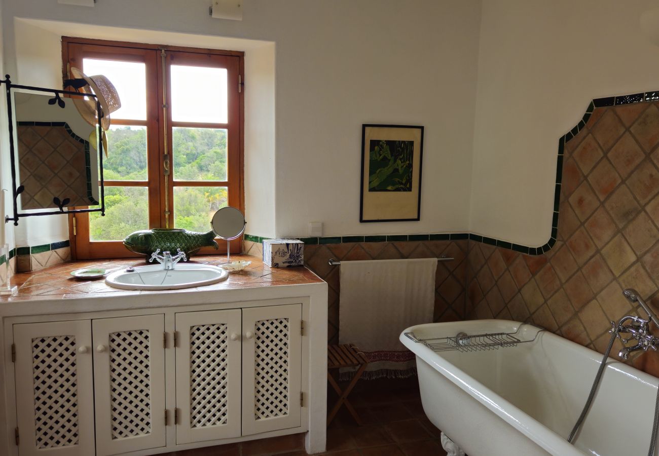 Charming bathroom featuring a Victorian-style soaking tub, rustic sink with a white wooden cabinet, and a wooden window overlooking lush greenery. Han