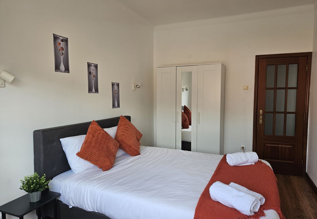 Spacious bedroom featuring a double bed, orange cushions, matching blanket, nightstand with decorative plant, and white wardrobe with mirror.