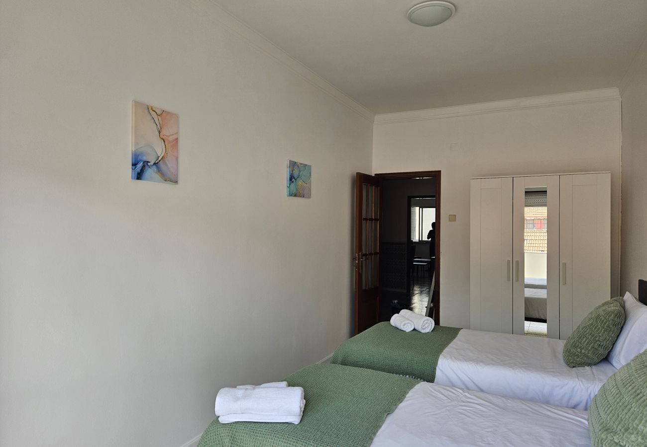 Spacious bedroom featuring two single beds, green bedding, matching cushions, a white wardrobe, and a wall adorned with abstract paintings.