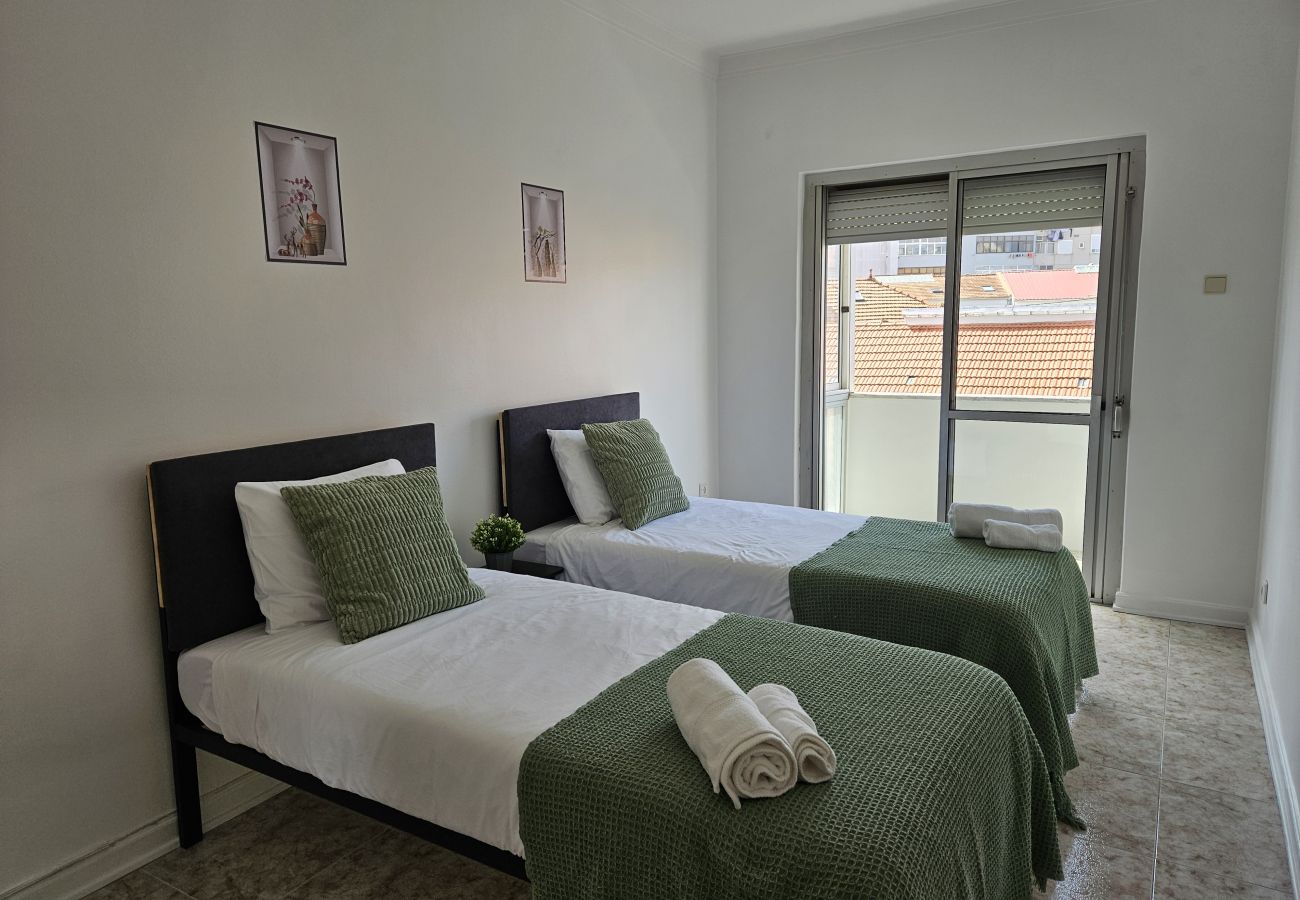 Bright bedroom featuring two single beds, green bedspreads, matching cushions, minimal decor, and a large window overlooking the city.