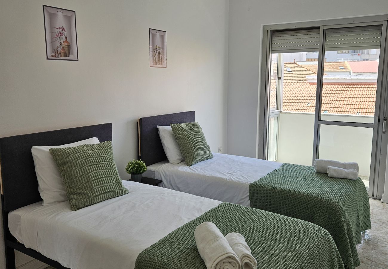 Bright bedroom featuring two single beds, dark headboards, white bedding with green blankets, minimalist decor, and a balcony overlooking the city.