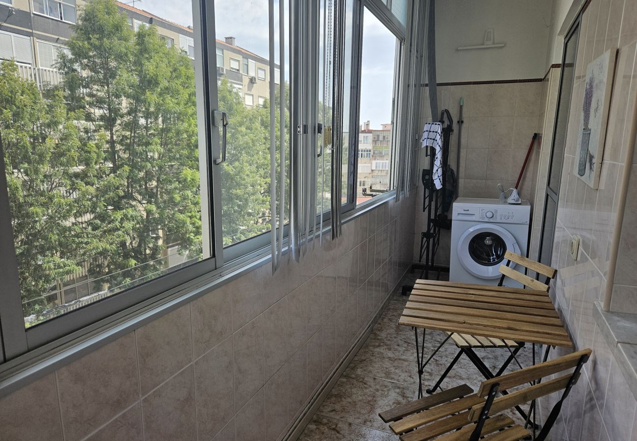 Enclosed balcony featuring a wooden folding table and chairs, a washing machine in the background, and a street view with trees and buildings.