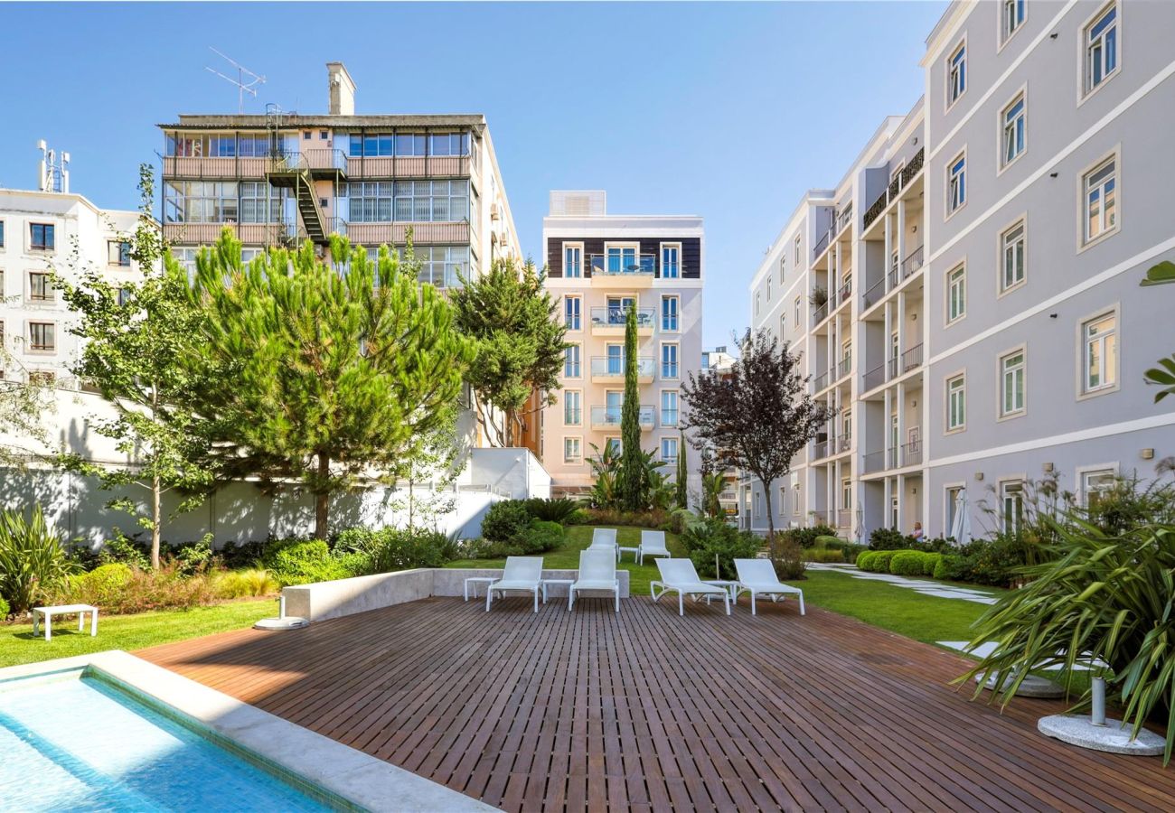  Wooden deck with white sun loungers next to the pool, surrounded by well-maintained gardens and modern buildings in the background.