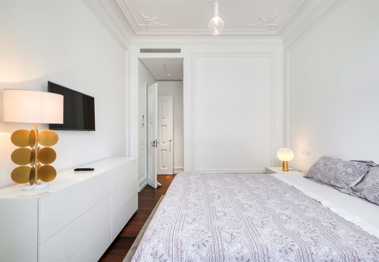 Spacious bedroom with double bed, lilac bedding, white dresser, wall-mounted TV, and sophisticated lighting.