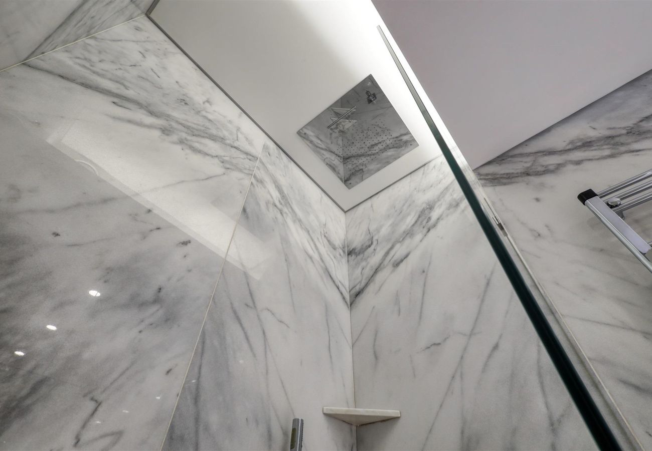 Detailed view of a ceiling-mounted shower, with walls covered in white marble and a minimalist shelf.
