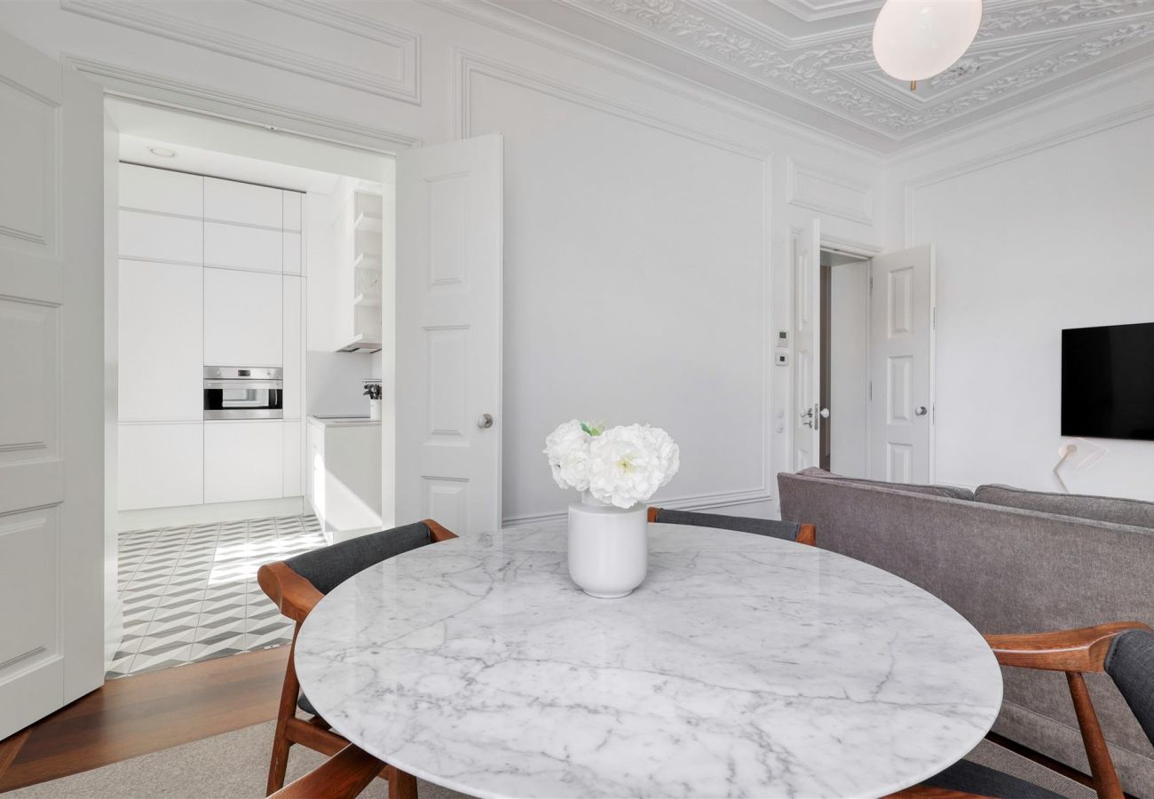 Round marble table with wooden chairs in a bright dining room, with access to a white kitchen in the background.